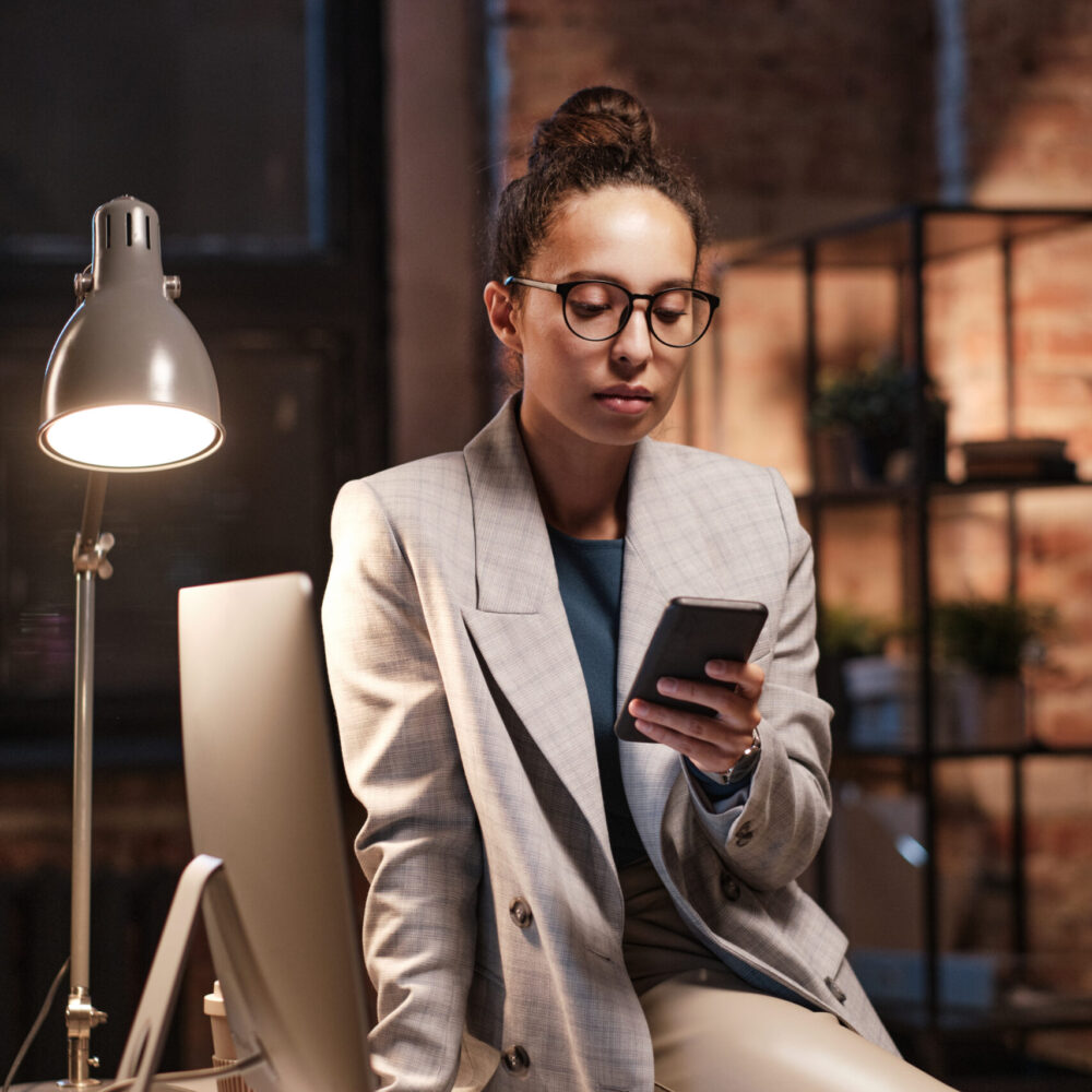 Serious young mixed race businesswoman in eyeglasses sitting on table with computer and scrolling through social media in dark office